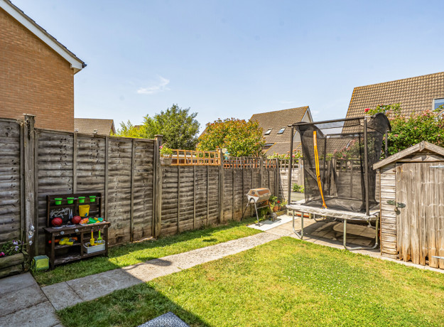 Garden with a lawn, paved path, fencing, trampoline and wooden shed.