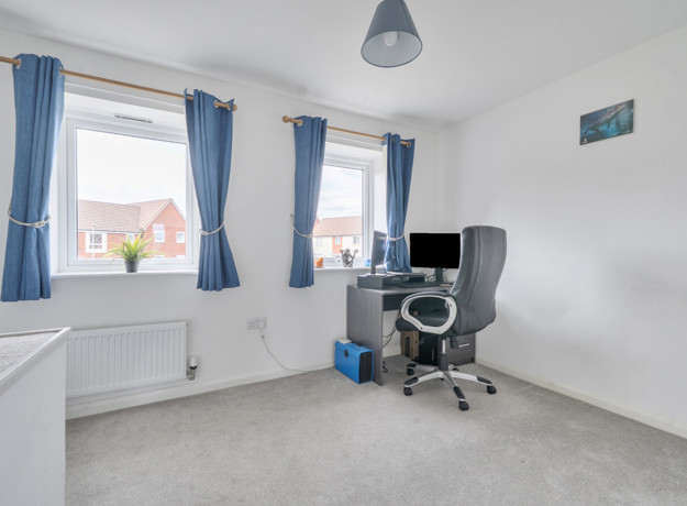 Room with desk, office chair, desktop screen and two windows with curtains.