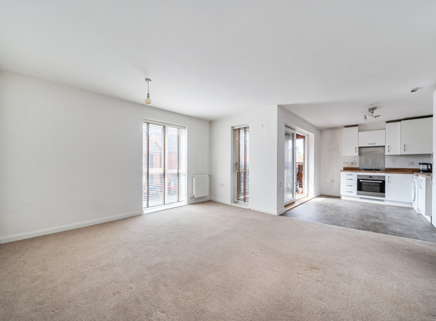 Open-plan living area with carpeted floor and kitchen in the background. (1)