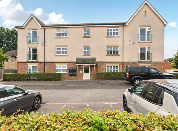 View of a residential apartment block with a central entrance, landscaped frontage, and parked cars along the road.