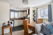 Dining area with wooden table beside compact kitchen and window with blinds.