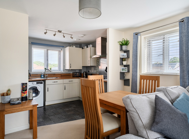 Dining area with wooden table beside compact kitchen and window with blinds.