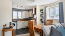 Dining area with wooden table beside compact kitchen and window with blinds.