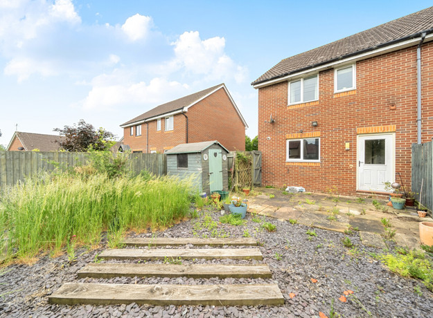Back garden with paving, grass patches, shed, and neighboring houses in view.