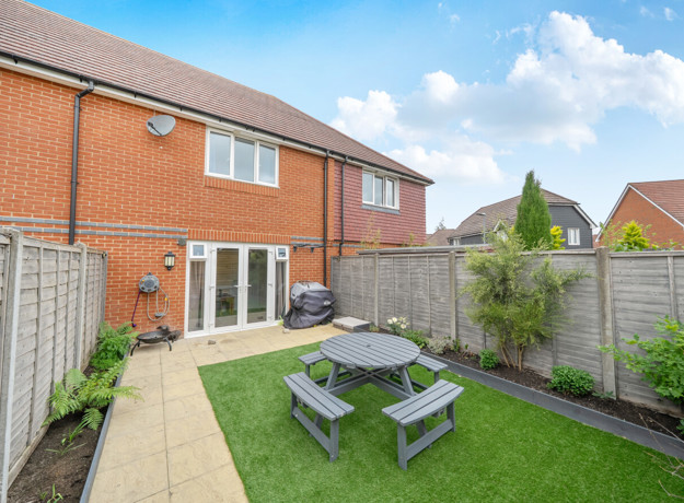 Back garden with patio, lawn, table and chairs enclosed by fencing.
