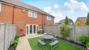 Back garden with patio, lawn, table and chairs enclosed by fencing.