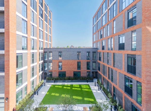 View into The Old Brewery residents' courtyard. Framed by The Old Brewery apartment blocks with large windows, featuring a rectangular landscaped green lawn, paved walkways, planted borders, and balcony railings under a clear blue sky.