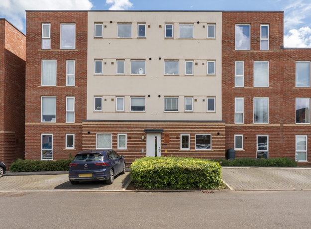 Apartment block with brick facade, cream panels, parked cars, and shrubs.