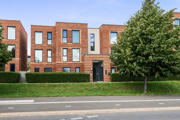 Modern brick apartment building with balconies, trees, and grass verge by the road.