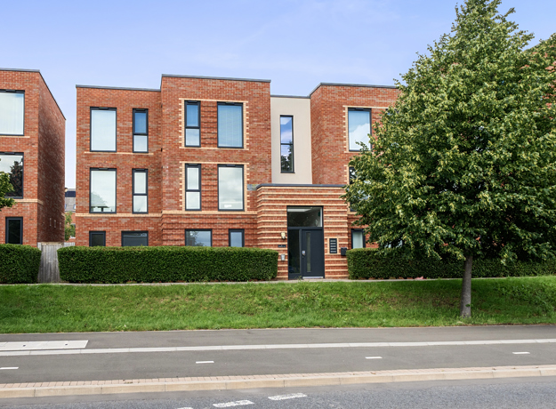 Modern brick apartment building with balconies, trees, and grass verge by the road.
