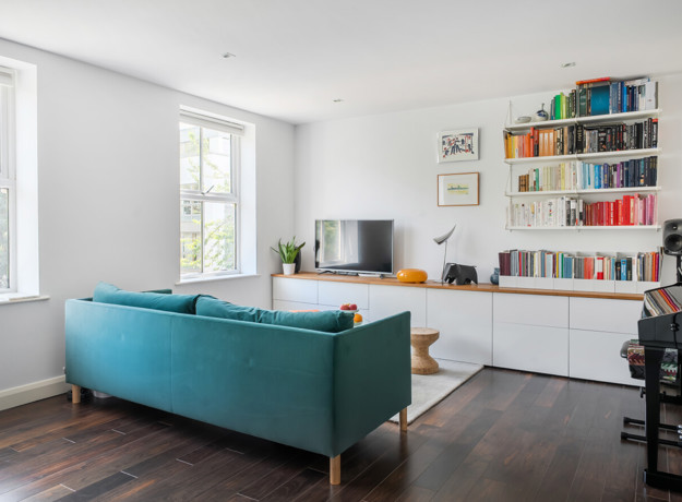 Living area with a teal sofa, wall-mounted shelves, piano and windows.