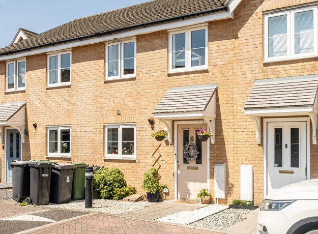Terraced brick houses with front doors, windows and wheelie bins along a paved street.